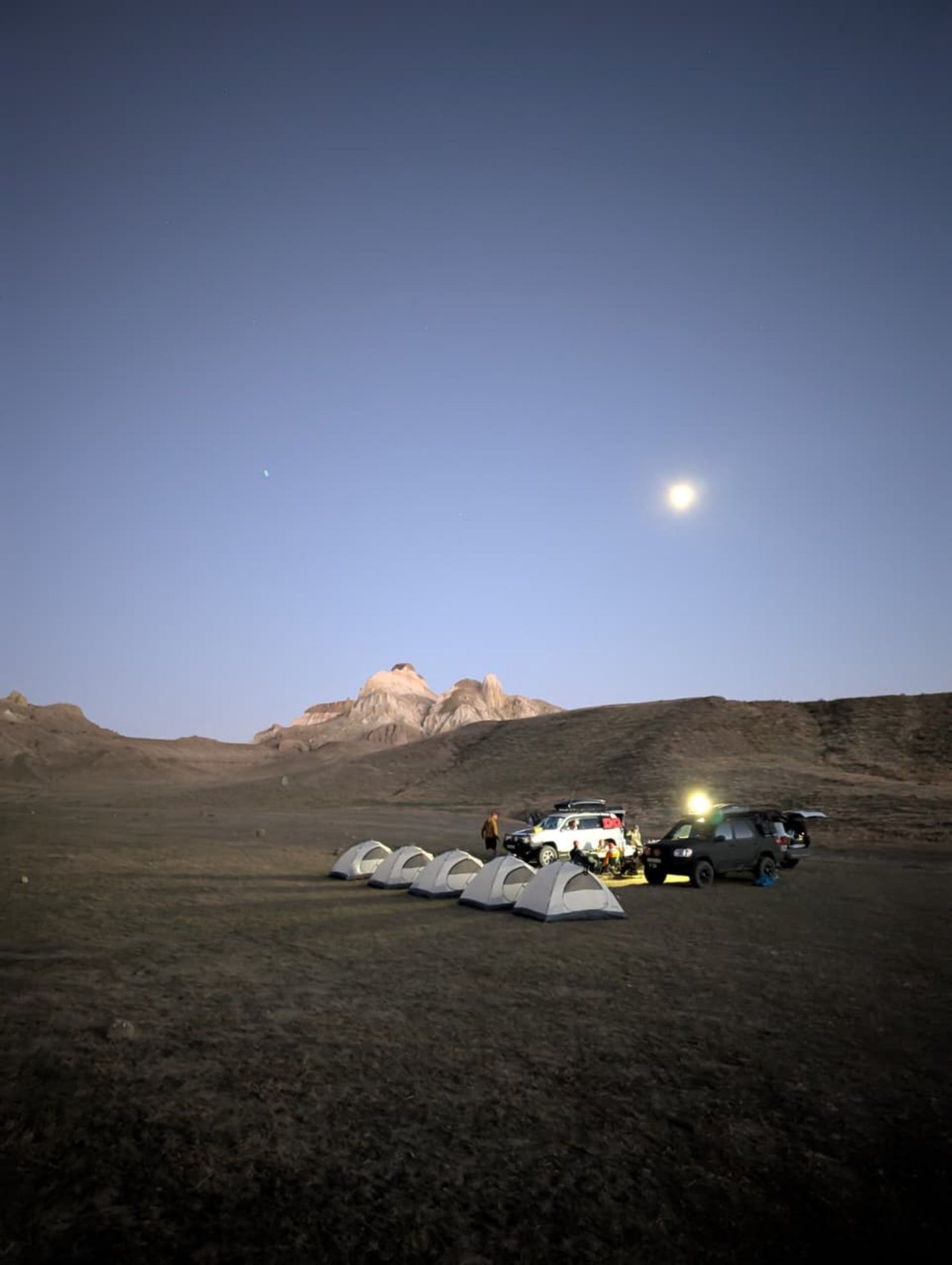 Nocny obóz w Mangystau – rząd namiotów rozstawionych w pustynnym krajobrazie, samochody terenowe oświetlone lampami i skaliste formacje górskie w tle przy świetle księżyca.